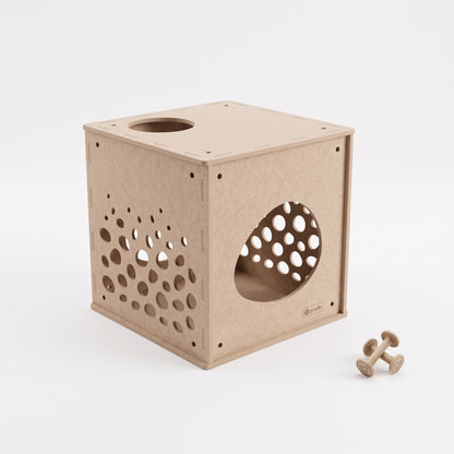 A light natural wood cube-shaped cat playhouse with circular entry and exit holes, shown on a clean white background with two small wooden toy dumbbells nearby.