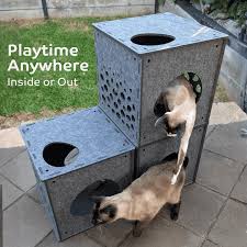A cat playhouse made of galvanized metal with multiple levels, holes, and perches, shown with two cats in an outdoor backyard setting. White text on the left reads 'Playtime Anywhere Inside or Out'.